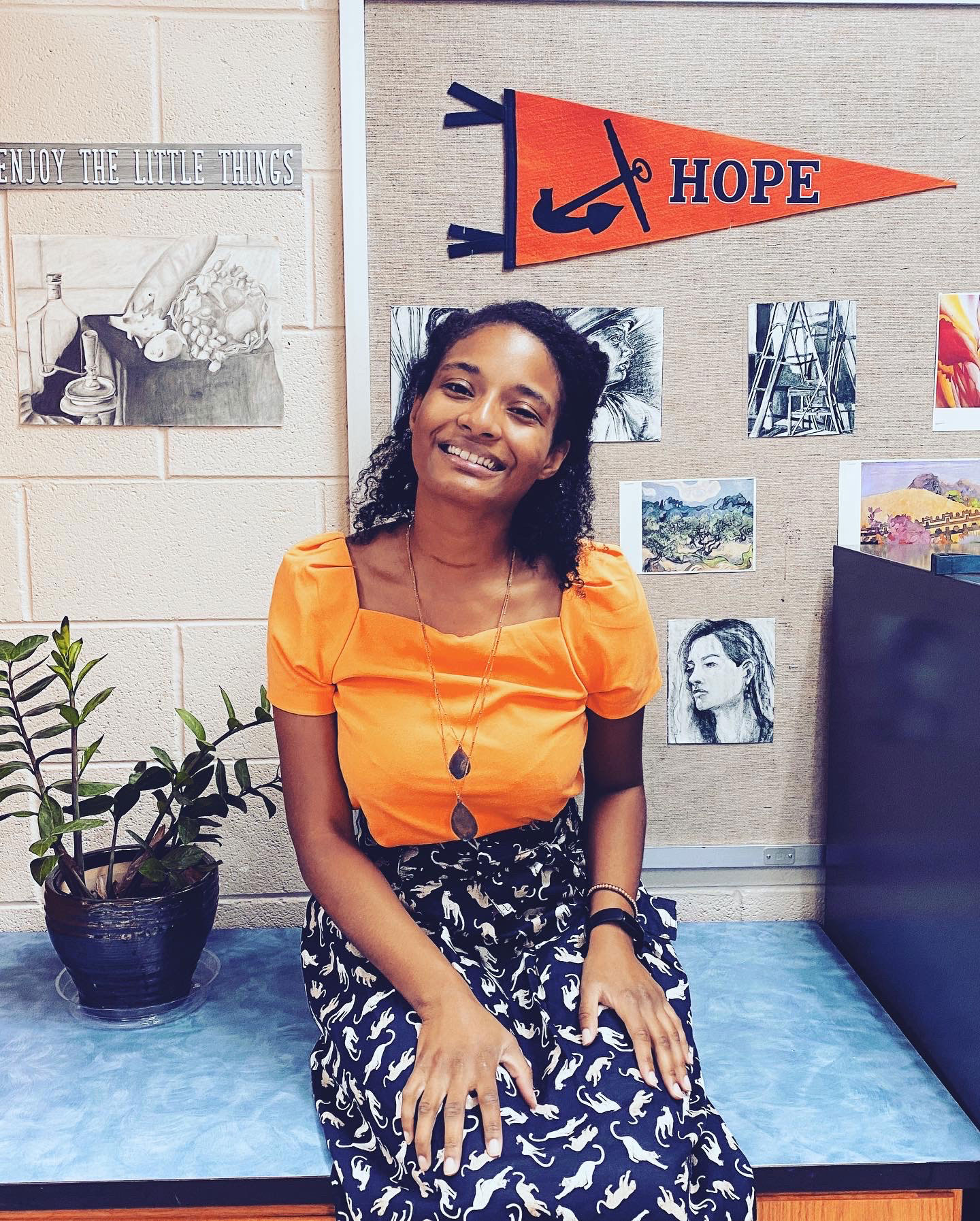 woman sitting on the edge of a desk, smiling, in front of student artwork