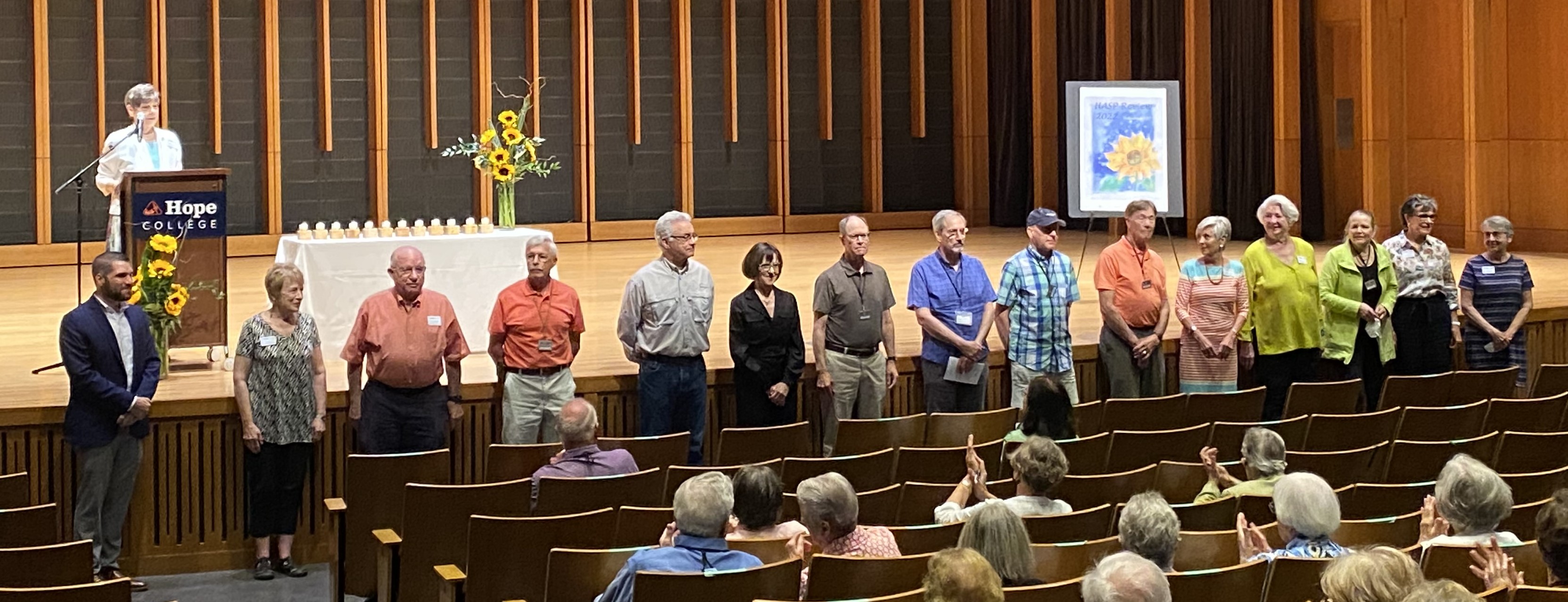 The 15 board of directors standing in front of a stage while a presenter speaks. 